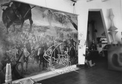 Fotografía en blanco y negro de una de las salas de a Casa Museo de Salvador Dalí en Portlligat. Se ve al artista bajo de una arco de entrada a la habitación, y una pintura de Dalí de grandes dimensiones en el suelo, apoyada en la pared de la derecha
